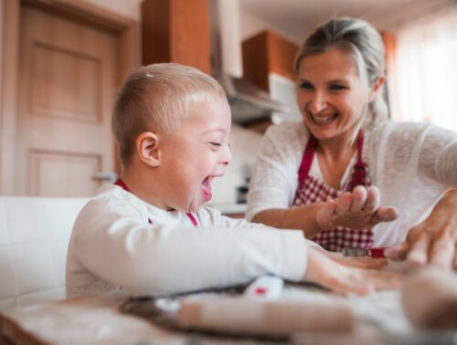 A laughing handicapped down syndrome child with his mother indoors baking.