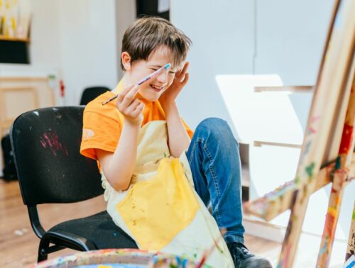Education and special child concept. Cute positive happy boy with down syndrome laughing while sitting in a chair and drawing with paintbrush indoor.